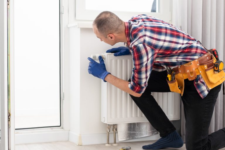 Man in work overalls using wrench while installing heating radiator in room. Young plumber installing heating system in apartment. Concept of radiator installation, plumbing works and home renovation. High quality photo